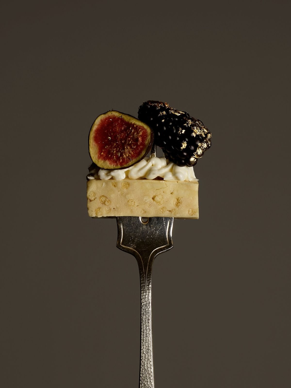 A single bite of cheese, cream, fig and a gold-leafed blackberry on a fork